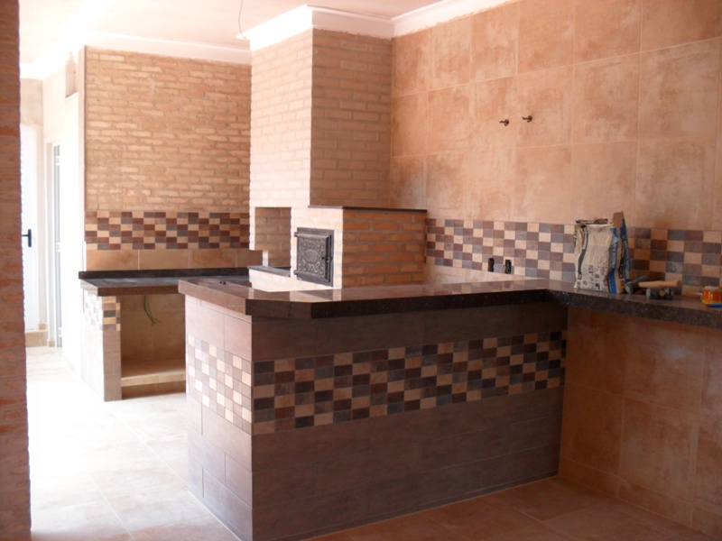 Cozinha externa integrada - Bancada em L com churrasqueira central, revestimento em tijolo e faixa decorativa em pastilha.
